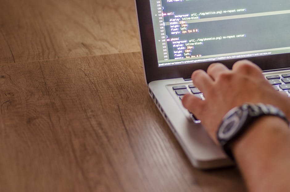A close-up view of a person's hand resting on a laptop keyboard, with the screen displaying lines of code or digital text. The laptop is placed on a wooden surface with a smooth, natural grain. The pe