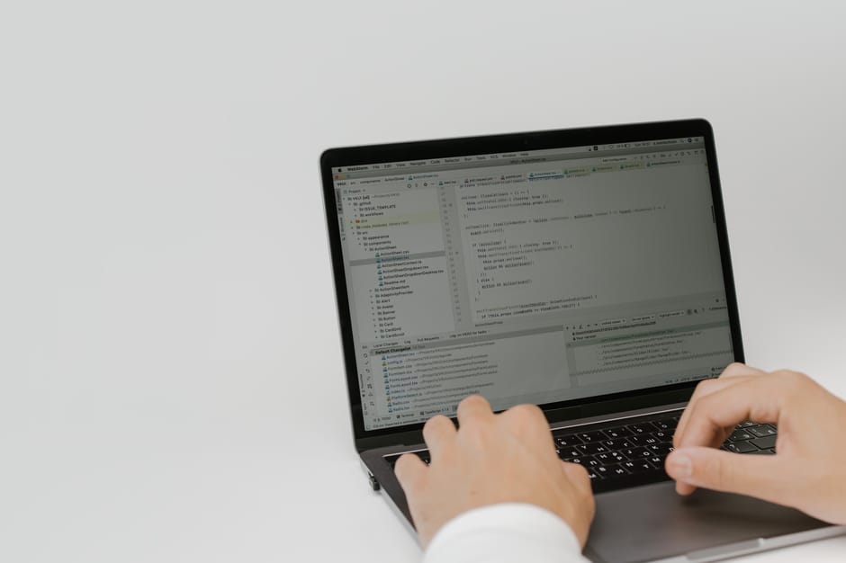 A person using a laptop to work on coding or programming, with the screen displaying a code editor filled with lines of code, folders, and options. The individual's hands are positioned over the keybo